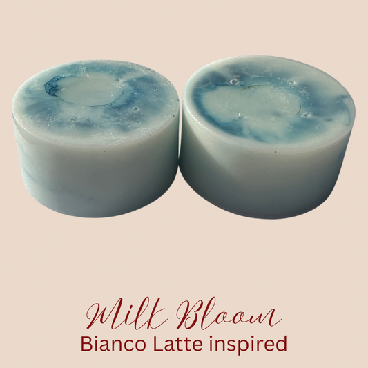 Two cylindrical light-blue wax melts with darker blue marbled swirls on top, displayed side-by-side on a beige background with the text 'Milk Bloom Bianco Latte inspired' underneath.